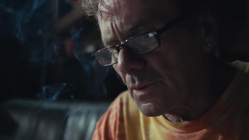 A pensive older man with glasses smokes a cigarette in a dimly lit room as smoke billows in slow motion