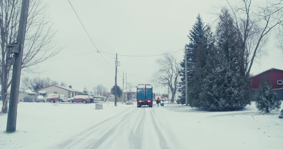 A sanitation worker collects recycling on a quiet, snow-covered residential street on a cold winter day