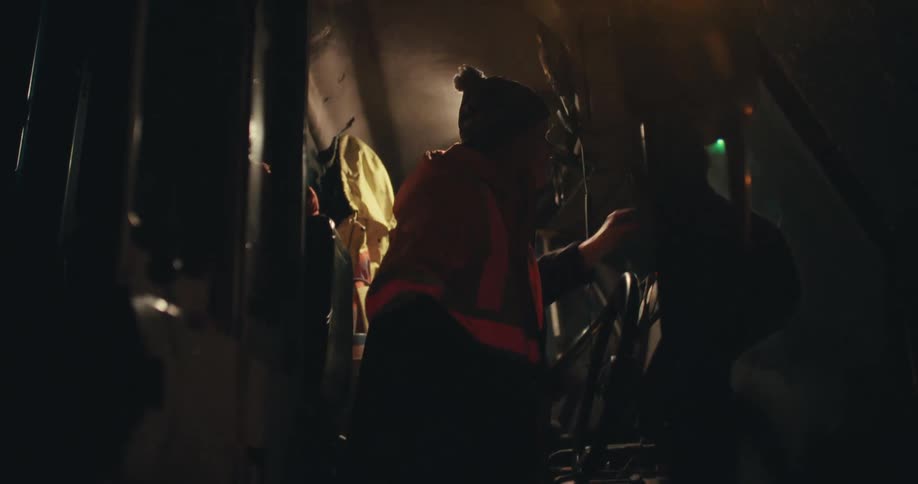 A municipal worker in a reflective vest operates a snowplow from inside the cab during a heavy nighttime snowstorm