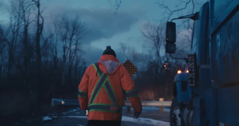 A sanitation worker in a bright orange jacket walks beside their truck on a cold, dark country road in slow motion