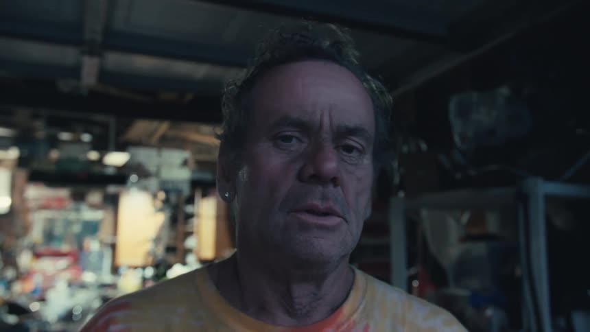 A weary-looking man in a tie-dye shirt takes a long drag from his cigarette in a dim, cluttered workshop