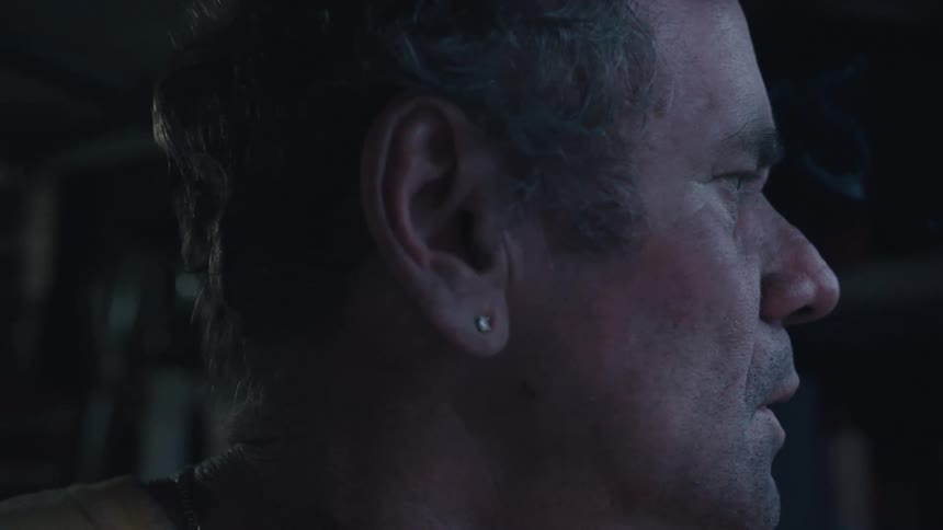 A moody, dramatic close-up of an older man's side profile as he thoughtfully takes a drag from a cigarette in a dark room