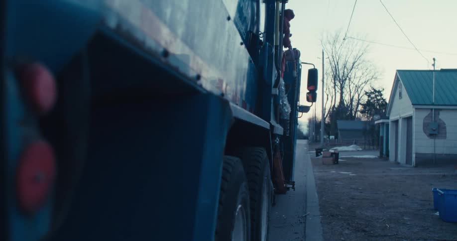 A sanitation worker collects recycling from a blue bin, gets back in his truck, and drives away down a quiet residential street