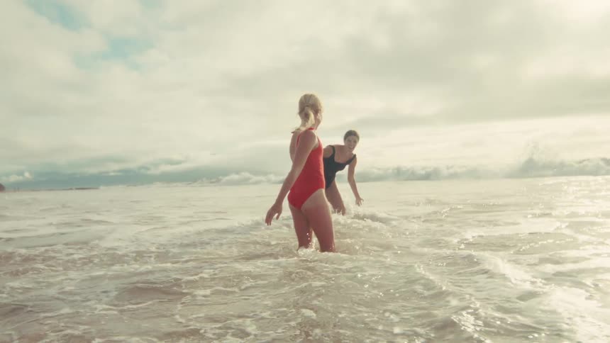 Two happy women in swimsuits playfully splash and wade in the ocean waves in slow motion