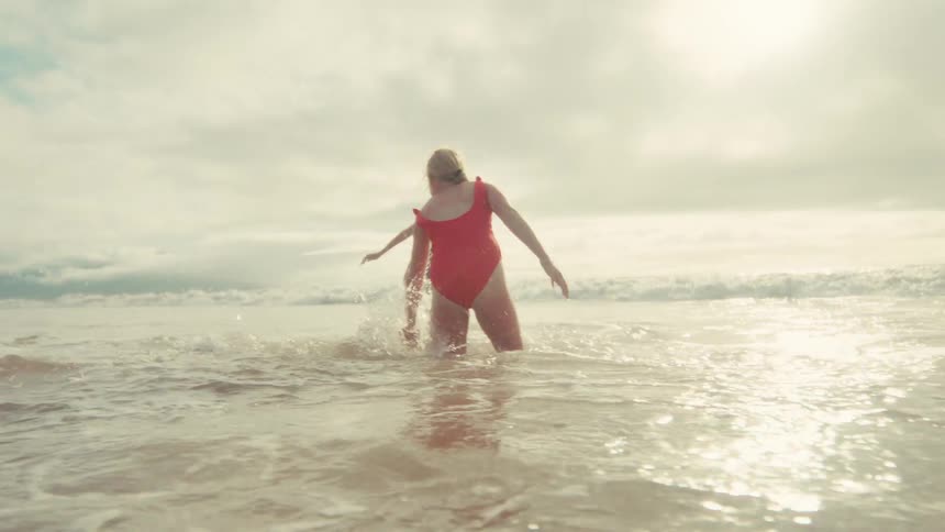 Two happy women in swimsuits playfully splash and wade in the ocean waves in slow motion