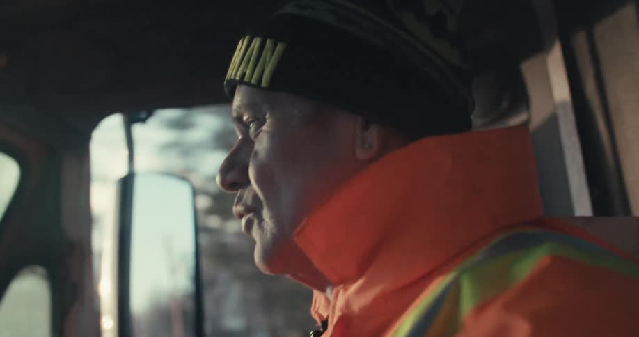A happy sanitation worker wearing a Batman beanie smiles while driving his truck on a sunny morning