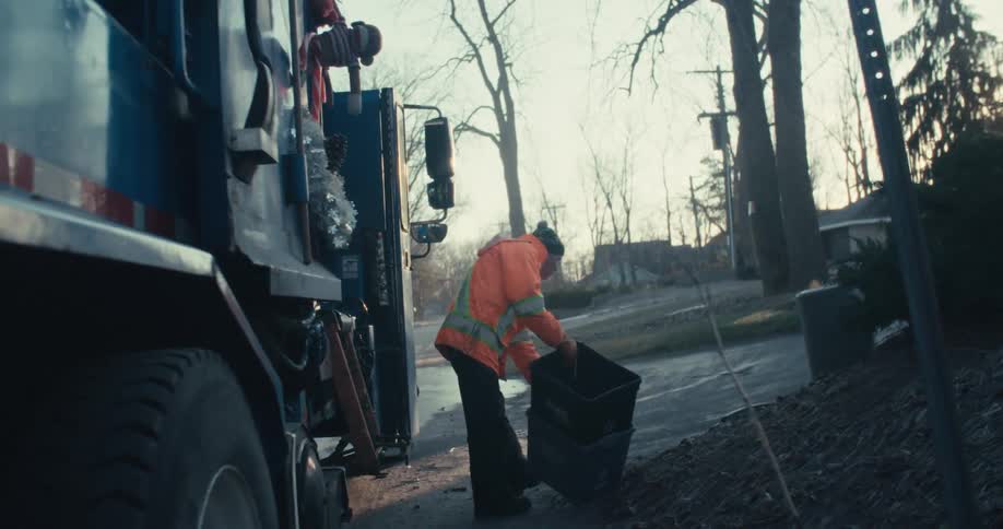 A sanitation worker finishes his pickup, returning an empty recycling bin to the side of the road before continuing his route in a large garbage truck