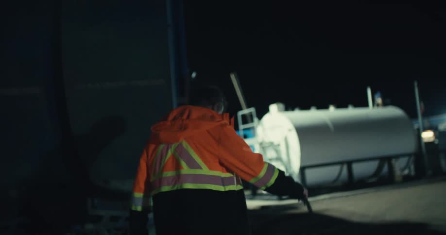A sanitation worker in a high-visibility jacket walks through the truck yard at night in slow motion