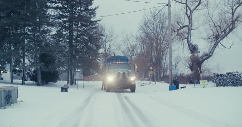 A recycling truck stops on a snowy residential road while a worker collects blue bins from the curbside on a cold winter day