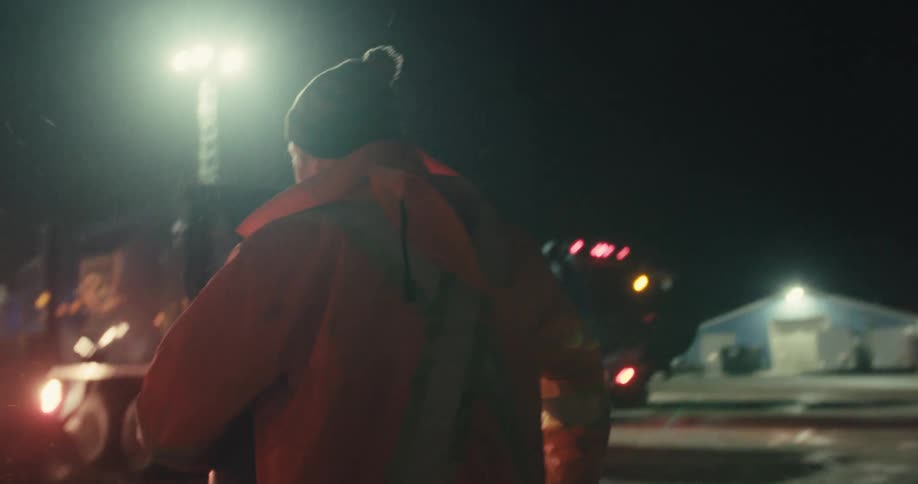 A sanitation worker in a high-visibility jacket walks through a snowy truck depot at night to begin their shift