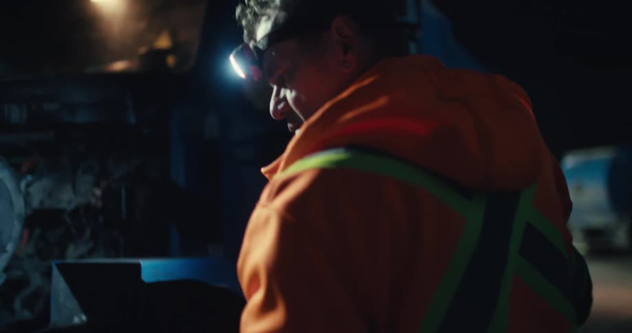 A mechanic in a safety vest uses a headlamp to inspect and work on a semi-truck's engine late at night