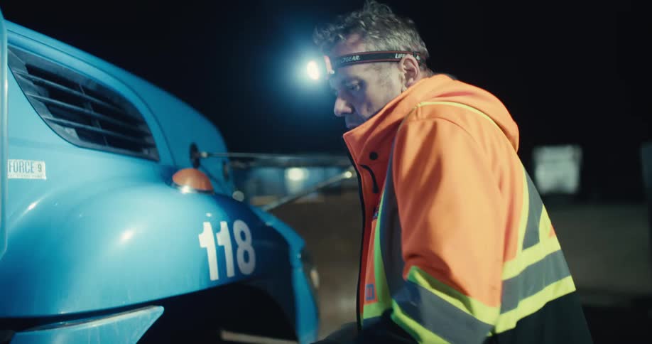A diligent worker in a safety jacket uses a headlamp to inspect his truck's tires during a night shift
