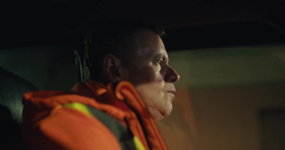A tired, blue-collar worker in a high-vis jacket drives his truck through the city at night, smoking a cigarette and drinking coffee