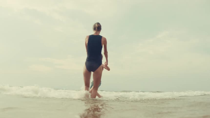 A mother and her young son playfully spin and splash in the ocean waves, holding hands and smiling in slow motion