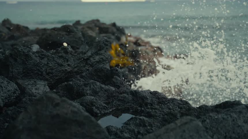 Slow-motion waves splash against a dark volcanic rock coastline as a cruise ship sits on the horizon