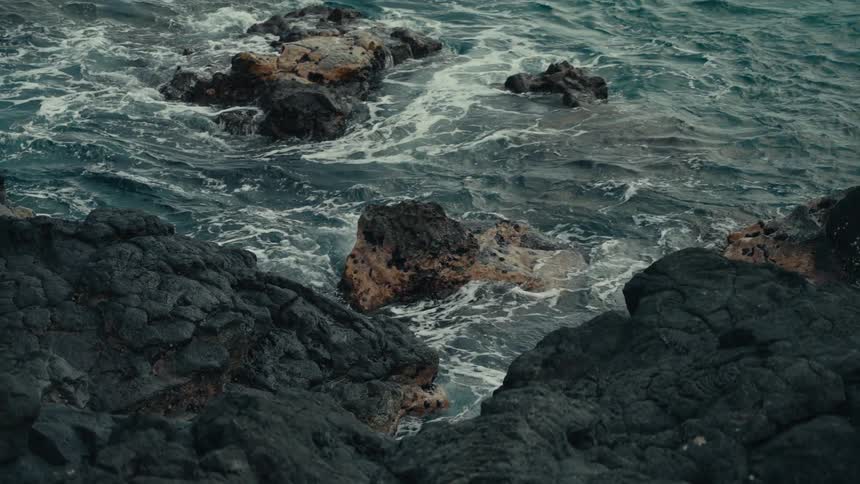A moody slow-motion shot captures the powerful yet mesmerizing churn of dark ocean waves against volcanic rocks on the shore