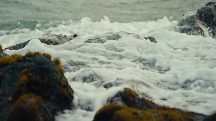 An ocean wave crashes in dramatic slow motion against mossy rocks, filling a tide pool with white foam