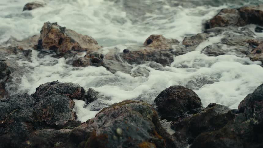 Ocean waves crash against rugged, mossy rocks in a dramatic slow-motion close-up