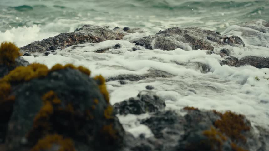A powerful ocean wave crashes over moss-covered rocks and tide pools in dramatic slow motion
