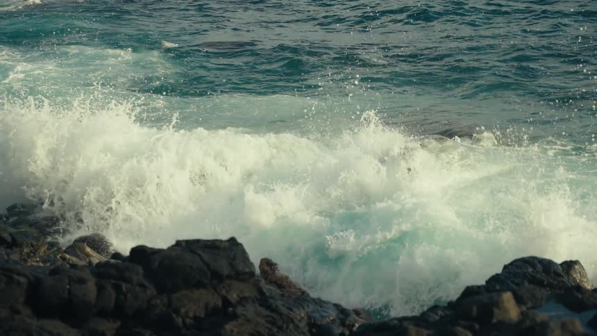 Powerful turquoise ocean waves crash against rugged volcanic rocks in dramatic slow motion