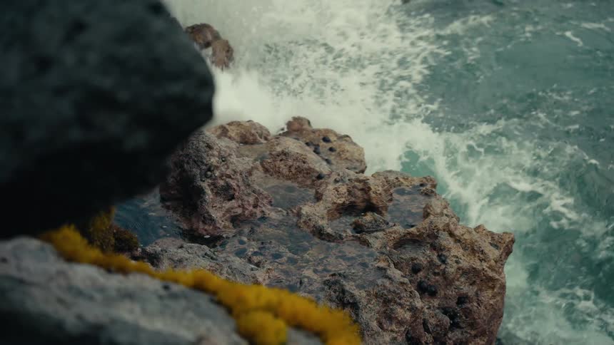 A powerful ocean wave crashes against rugged volcanic rocks draped with a yellow lei in dramatic slow motion