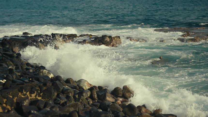 Dramatic slow-motion footage shows turquoise ocean waves crashing against a rugged, rocky shore