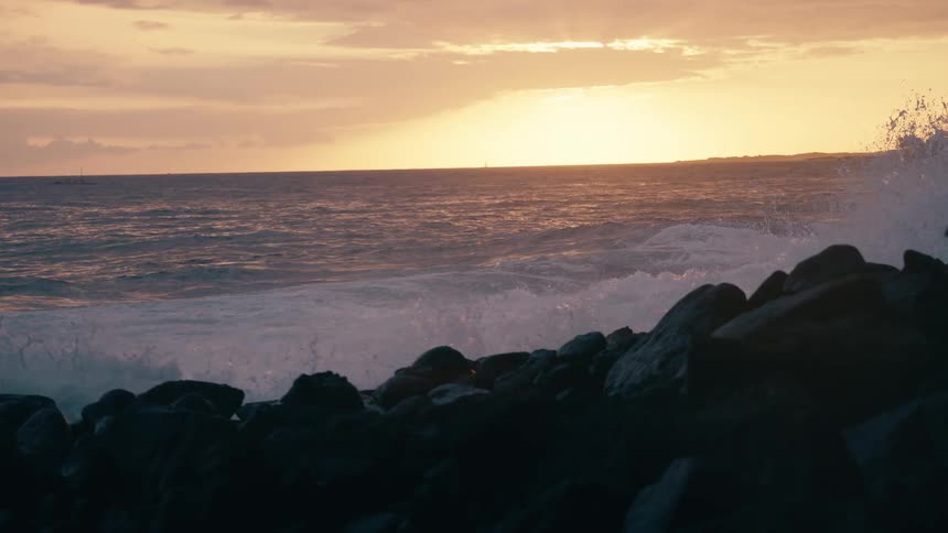 Ocean waves crash against a rocky shoreline in dramatic slow motion during a vibrant sunset