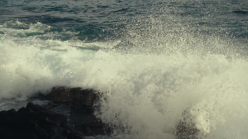 A powerful turquoise wave curls and crashes against a rocky shore in slow motion
