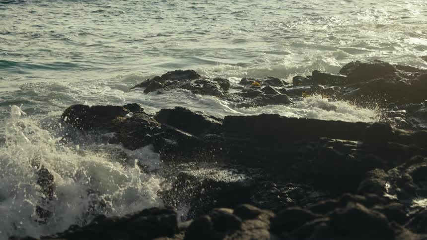Ocean waves crash over dark rocks in slow motion, illuminated by the warm light of the sun