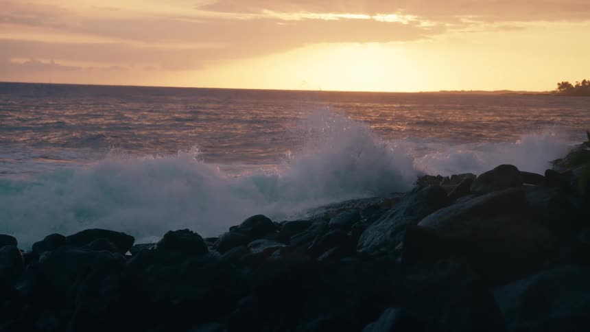 Powerful ocean waves crash in slow motion against a rocky shore as the golden sun sets on the horizon