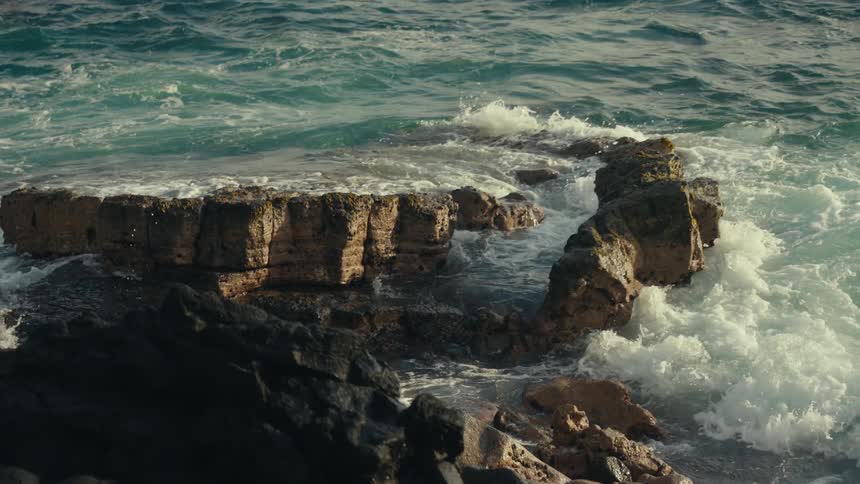 Slow motion footage shows turquoise ocean waves crashing against a rugged, rocky shoreline