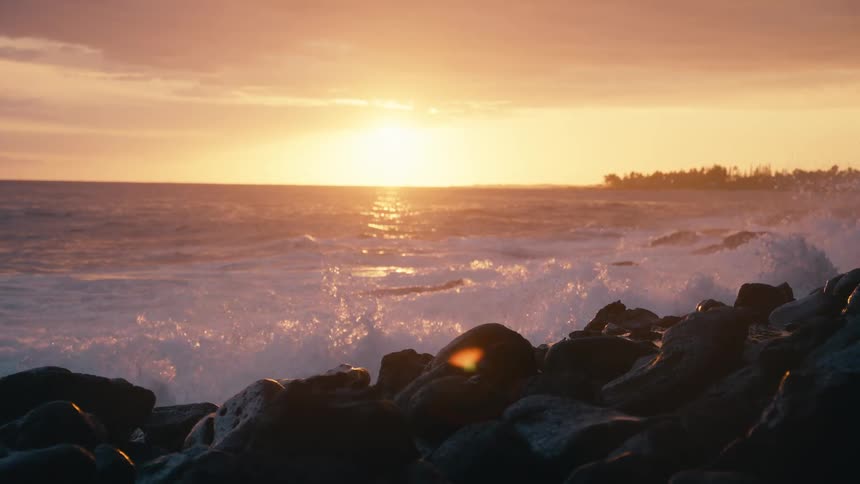 A powerful ocean wave crashes onto a rocky beach in stunning slow motion as the sun sets on the horizon
