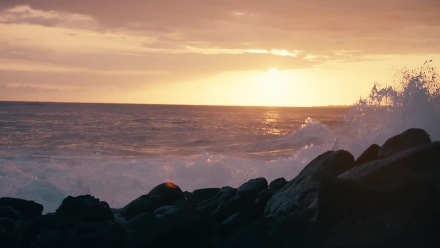 Slow motion waves crash against a rocky shoreline during a vibrant golden sunset over the ocean