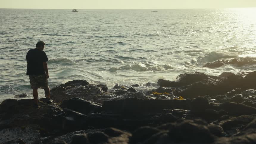 A solitary man stands on a rocky coastline, gazing at the ocean as waves crash against the shore in slow motion during a hazy sunset