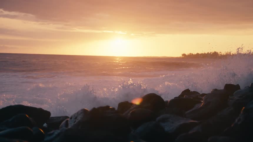 Golden hour sun sets over the ocean as waves majestically crash onto the rocky coastline in slow motion