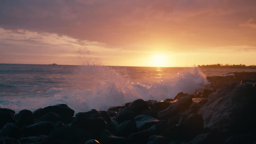 A powerful ocean wave crashes onto a rocky shoreline in stunning slow motion as the golden sun sets on the horizon