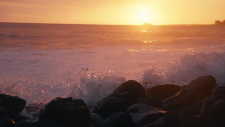 Dramatic super slow motion of powerful ocean waves crashing over a rocky shoreline during a golden sunset