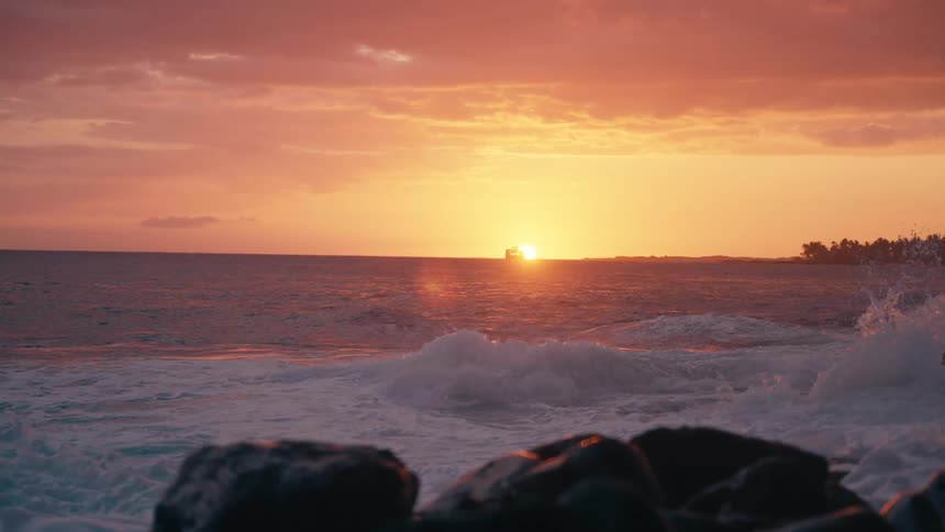 Slow motion of powerful ocean waves crashing against rocks during a vibrant sunset with a ship on the horizon