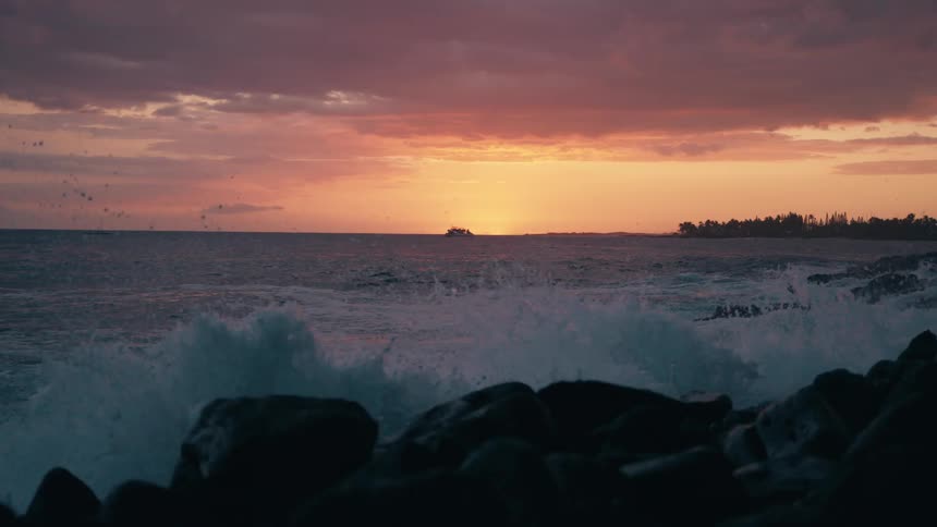Slow-motion footage shows vibrant turquoise waves crashing against a rocky shoreline as the sun sets over the ocean