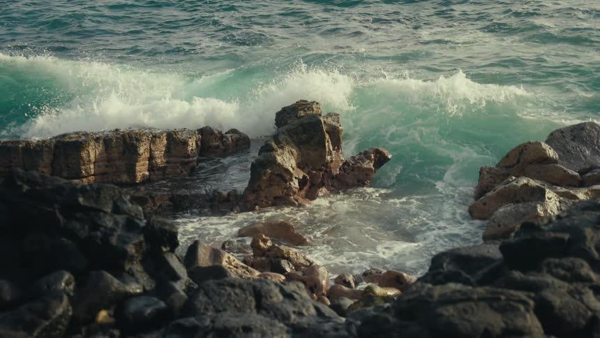 A powerful turquoise ocean wave rolls in and crashes against a rugged, dark rocky shoreline in dramatic slow motion
