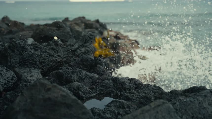 Close-up slow-motion shot of an ocean wave breaking over a jagged lava rock shore
