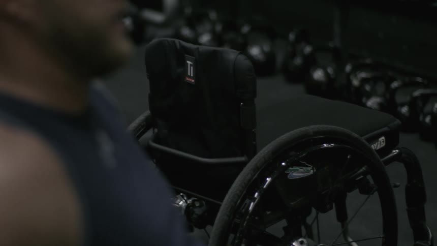 A wheelchair sits nearby a man who is lifting weights in a gym