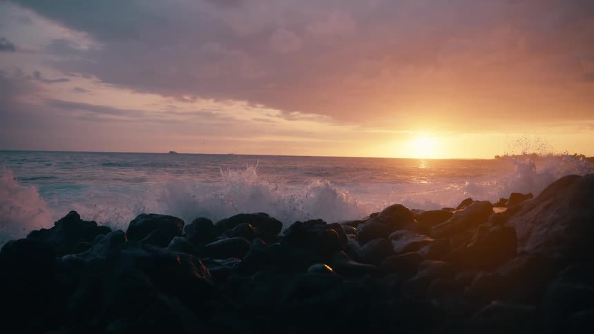 Dramatic slow-motion ocean waves crash on a rocky shore during a beautiful golden sunset