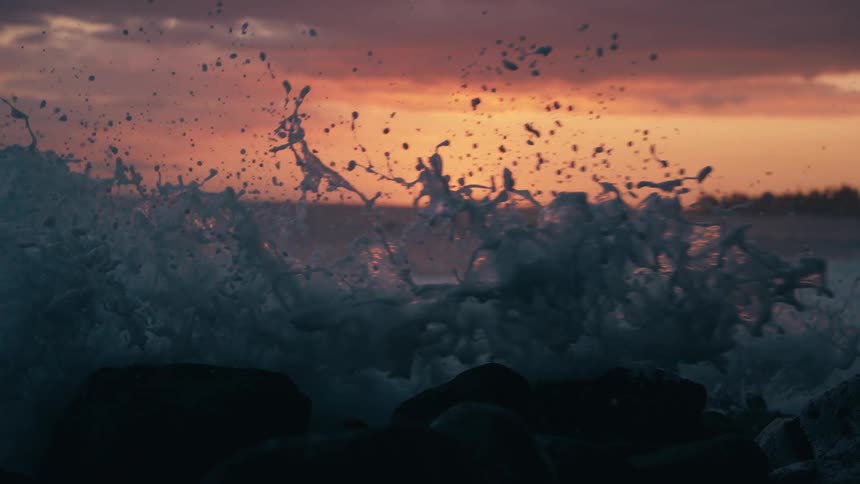 A powerful ocean wave crashes against a rocky shore in dramatic super slow motion during a vibrant sunset