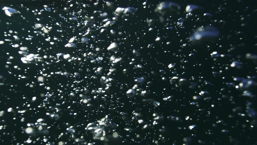 The camera slowly submerges into the ocean, revealing a magical display of sunlit bubbles rising through the dark water