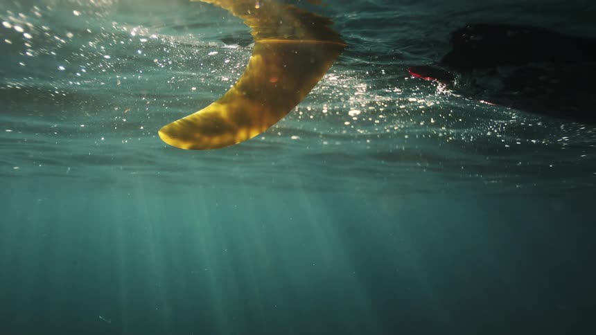 The yellow fin of a surfboard floats on the ocean surface, seen from below as beautiful sun rays shine through the water
