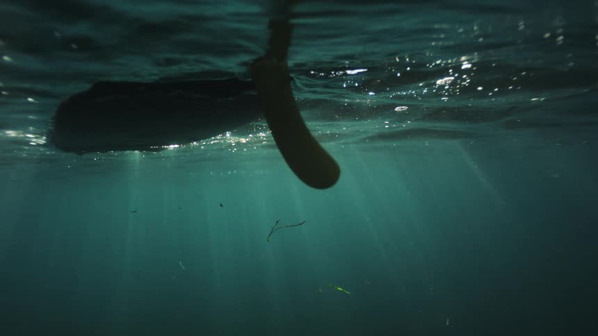 An ethereal underwater perspective captures a surfboard fin suspended in the ocean as beautiful sunbeams pierce the water's surface