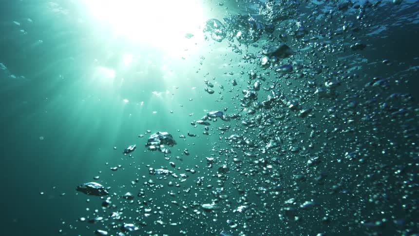 A slow-motion underwater view shows countless bubbles rising through tranquil turquoise water toward the sun's brilliant light
