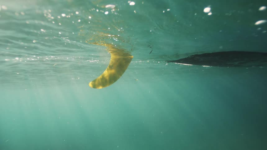 An underwater view shows the fin of a surfboard floating in the tranquil ocean as sun rays pierce through the water's surface