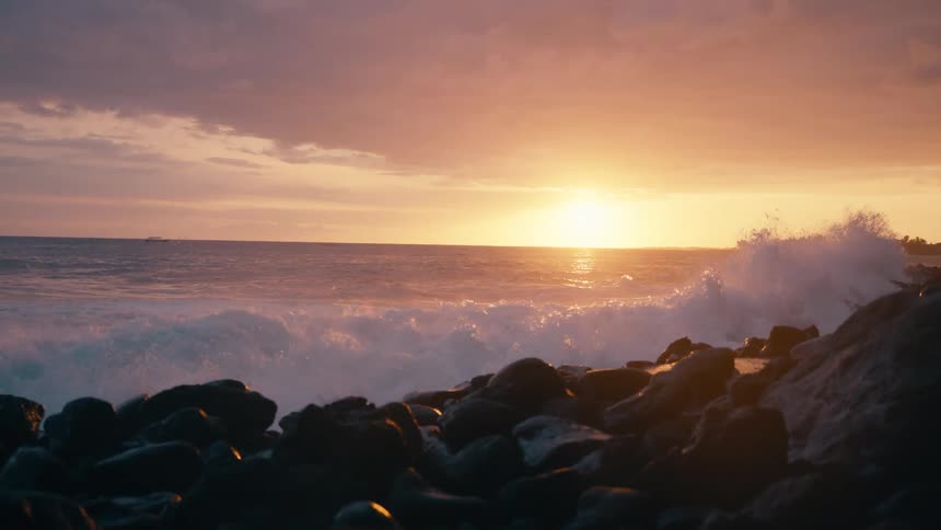 A powerful ocean wave crashes against a rocky shore in dramatic slow motion during a beautiful golden sunset
